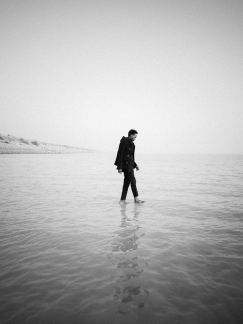 A man walks barefoot along the calm seaside in a peaceful black and white scene.