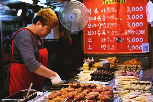 A street food vendor prepares Korean skewers at an outdoor market with a vibrant menu in the background.