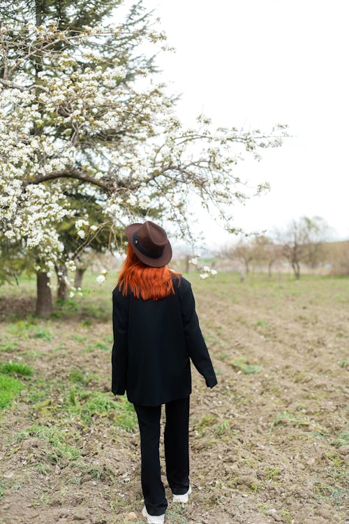 A woman with red hair and a hat walks through a field
