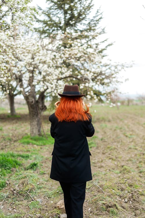 A woman with red hair and a hat walks through an orchard
