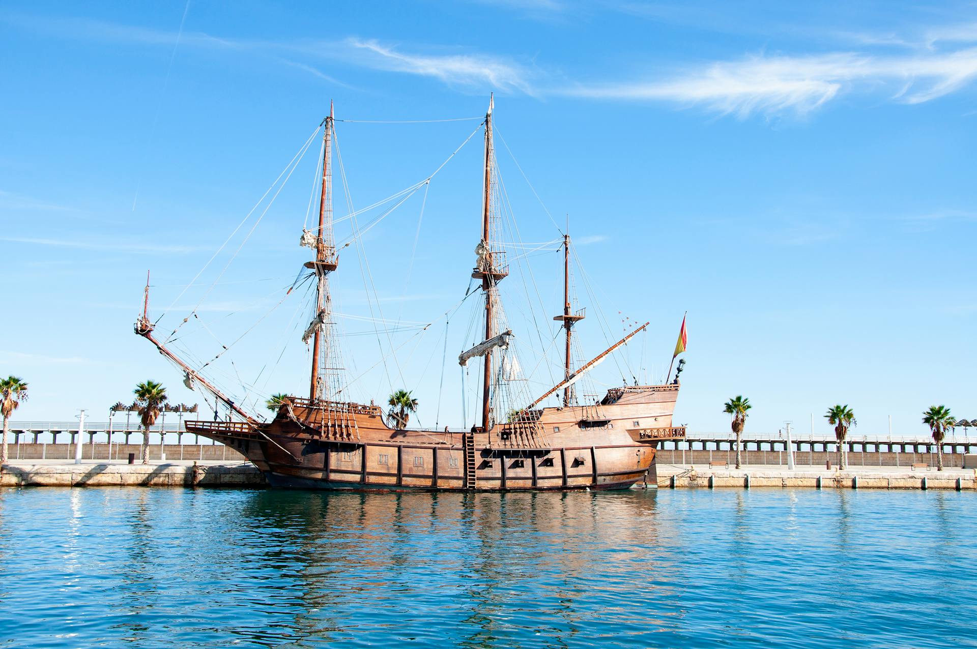 Wooden galleon docked at a sunny Mediterranean harbor with palm trees and blue skies.