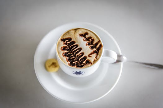 Top view of cappuccino with intricate chocolate design, next to a cookie on a clean, white table setting.