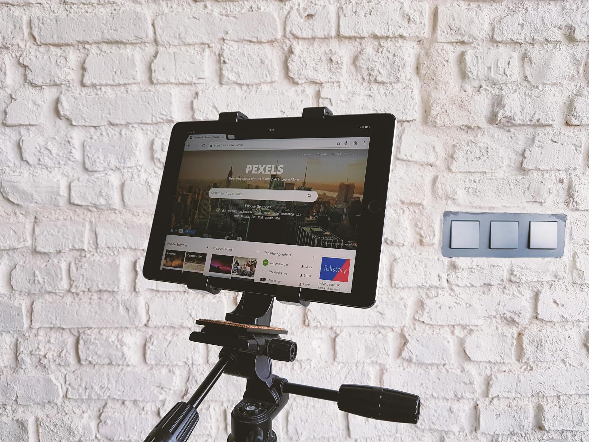 Tablet mounted on a tripod, displaying a website, set against a white brick wall indoors.