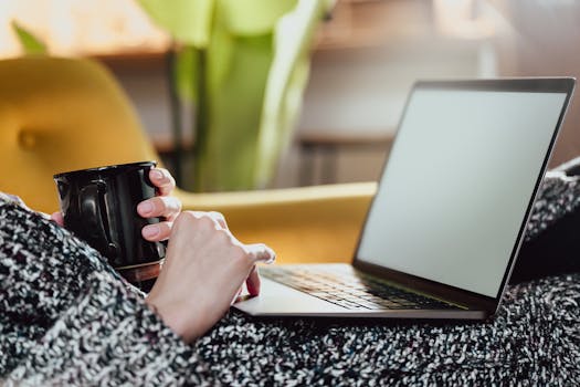 Cozy setting with a woman holding a coffee cup and using a laptop on a sofa.