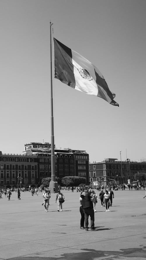 A black and white photo of people walking around a plaza with a mexican flag