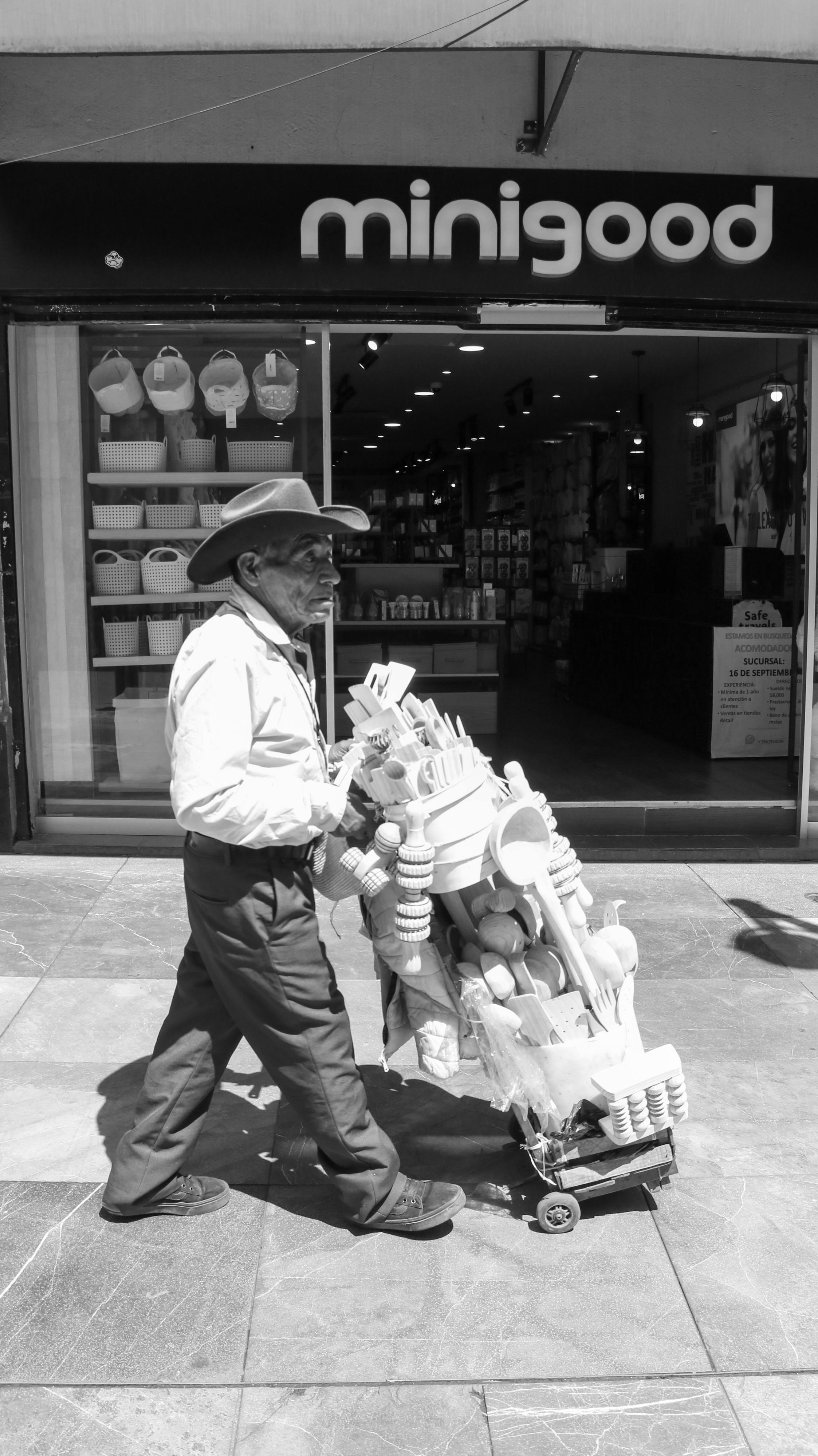 Black and white image of a street vendor pulling a trolley past Minigood store.