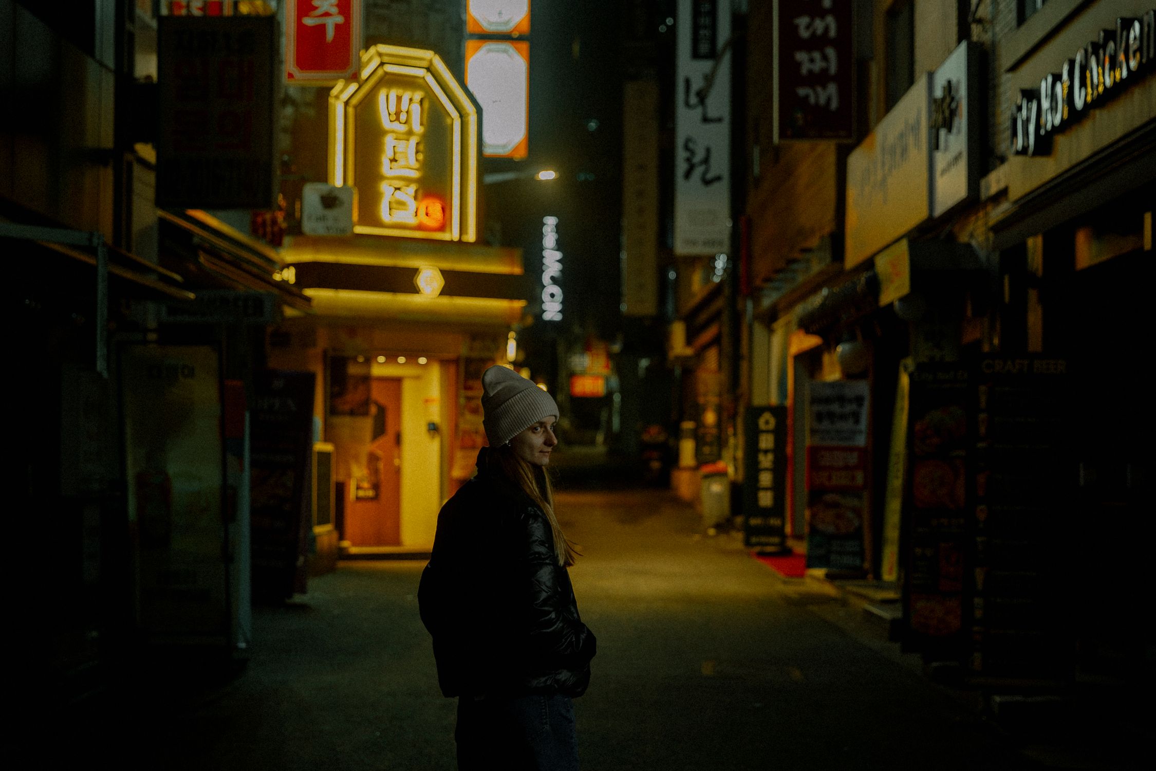 Woman in beanie and jacket standing in dimly lit urban alley in Seoul at night