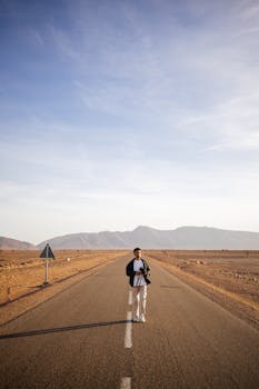 A solo traveler walks along a deserted road with mountains in the background under a clear sky.