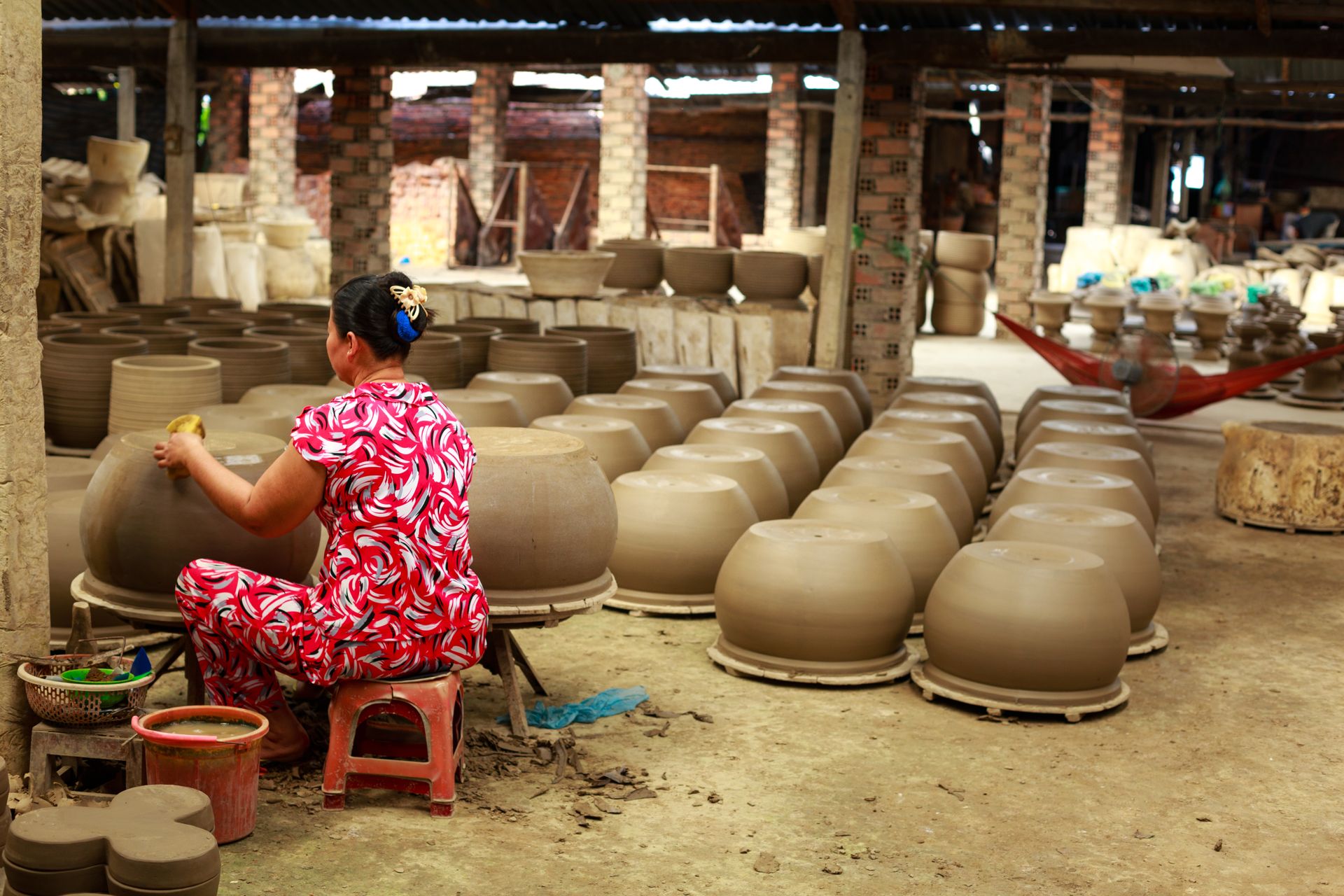 An artisan woman creates pottery in a traditional workshop, surrounded by clay pots and earthenware pieces.