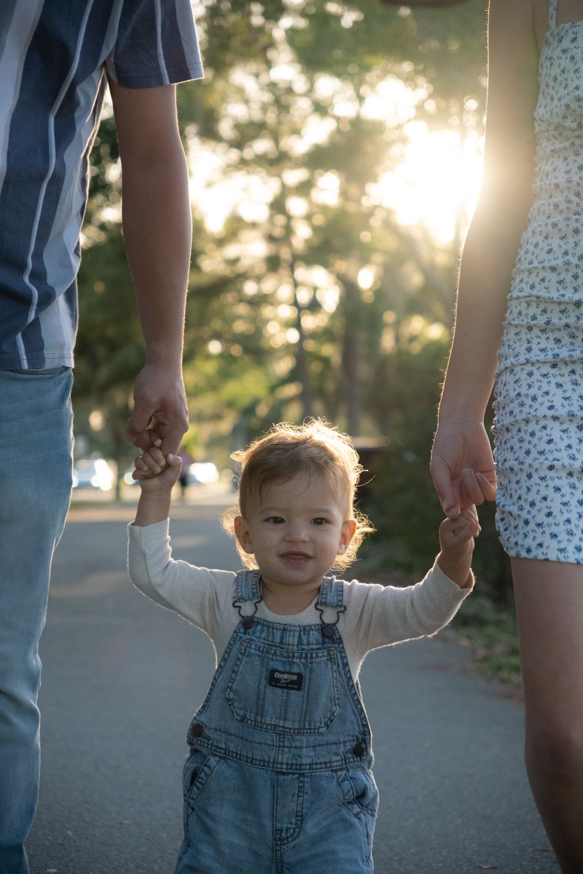 A young child holding hands with their parents on a sunny park path