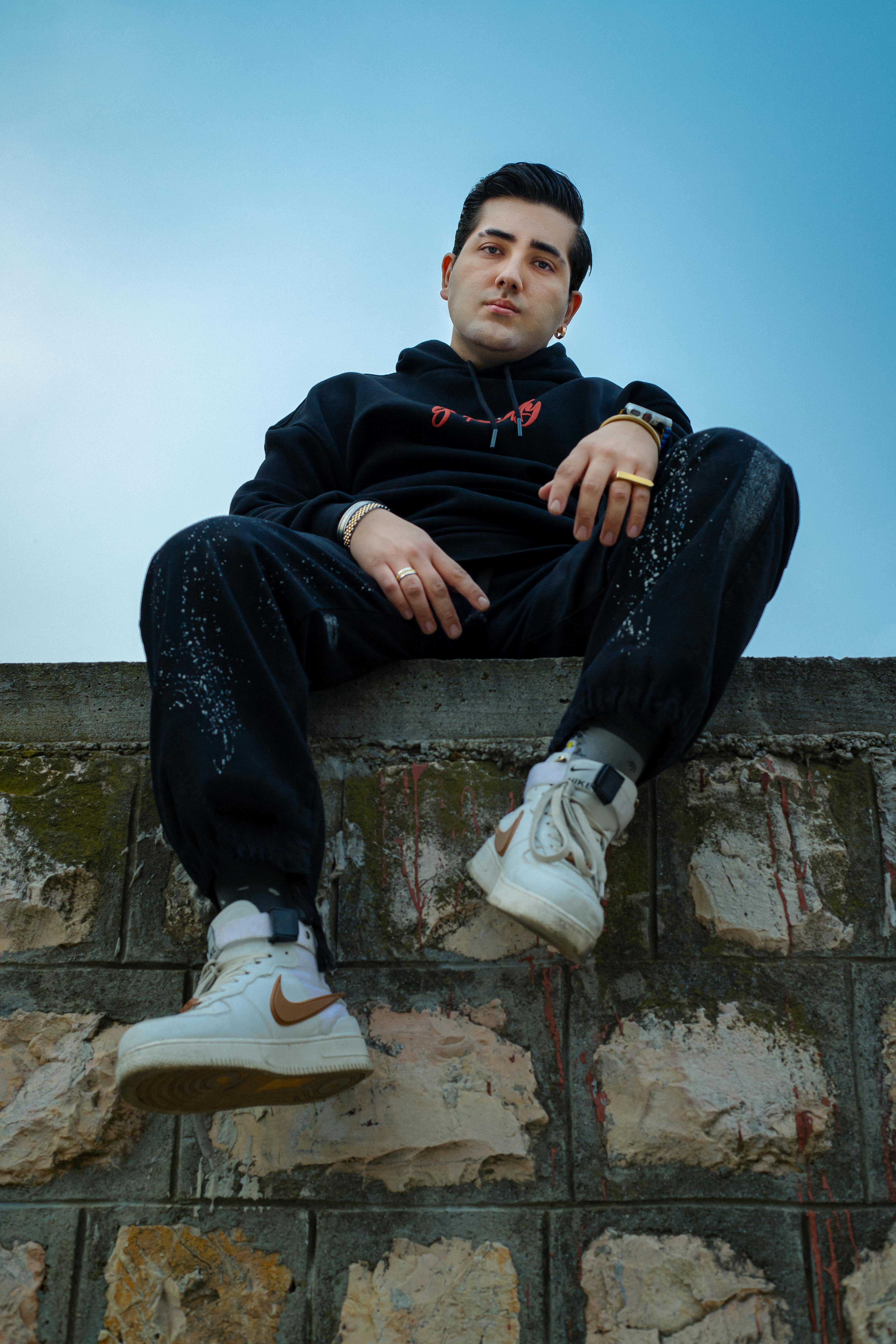 Stylish young man in black hoodie and sneakers sitting on a wall outdoors with clear blue sky backdrop.