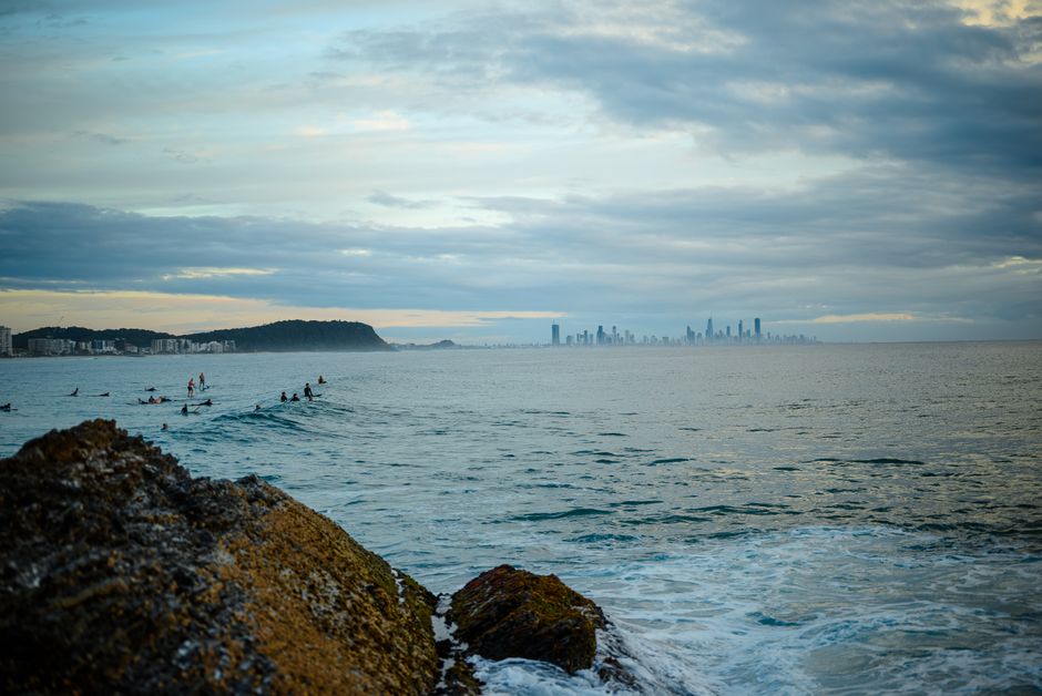 Surfers catching waves with a view of Gold Coast skyline from Currumbin Beach during sunset.