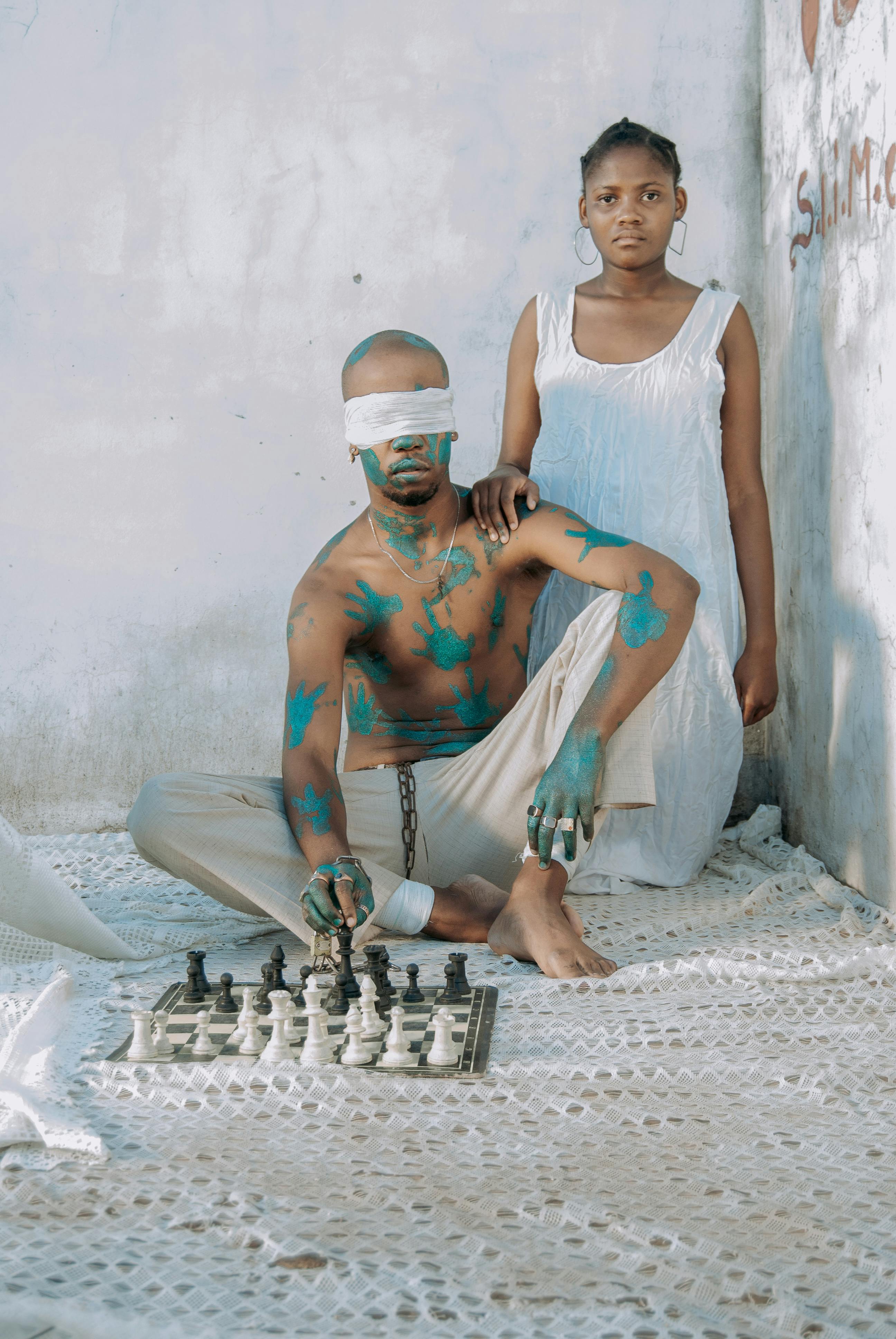 man playing chess in a blindfold and with green handprints on his skin accompanied by a woman