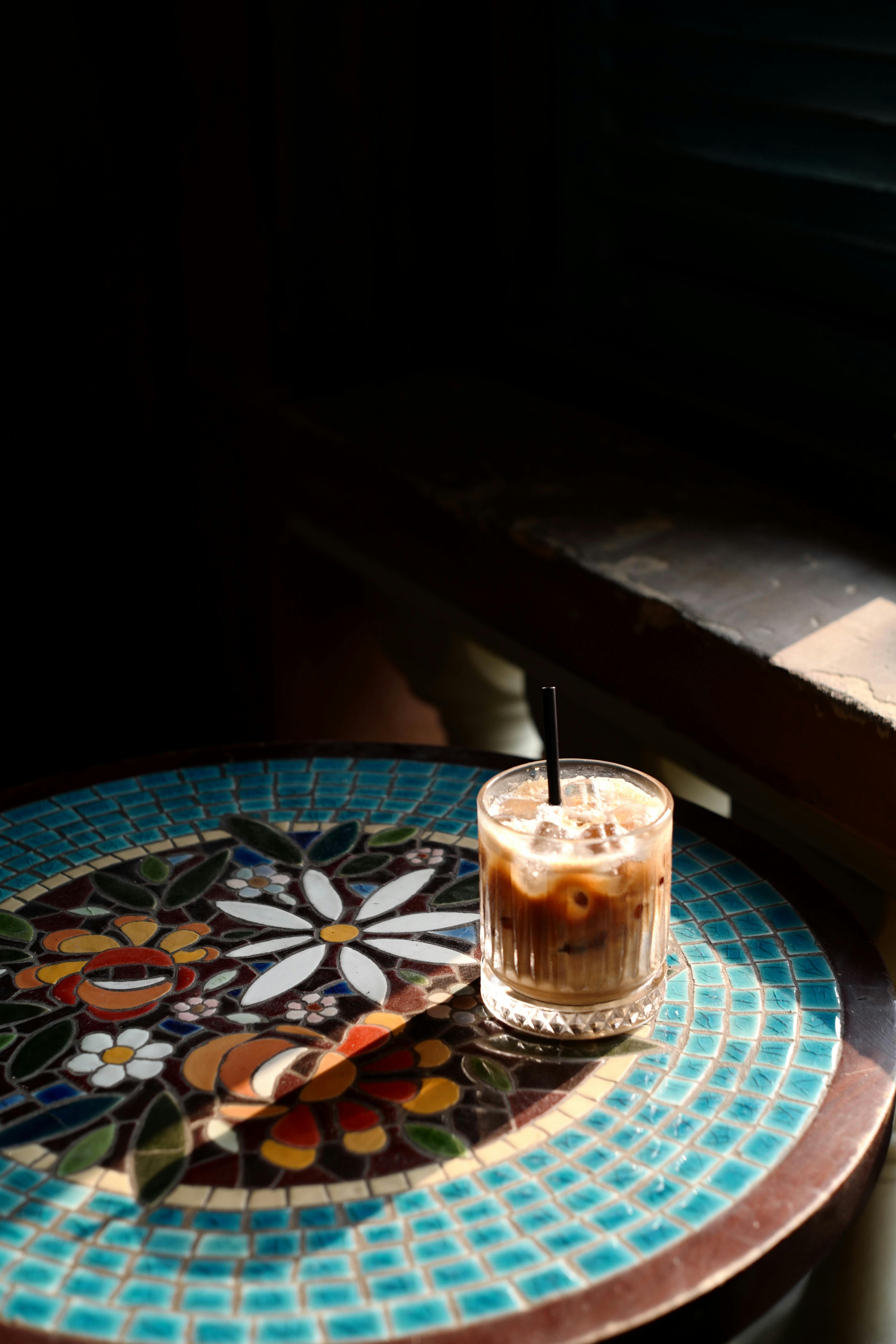 A glass of iced coffee with a straw sits on a vibrant mosaic table in natural light.