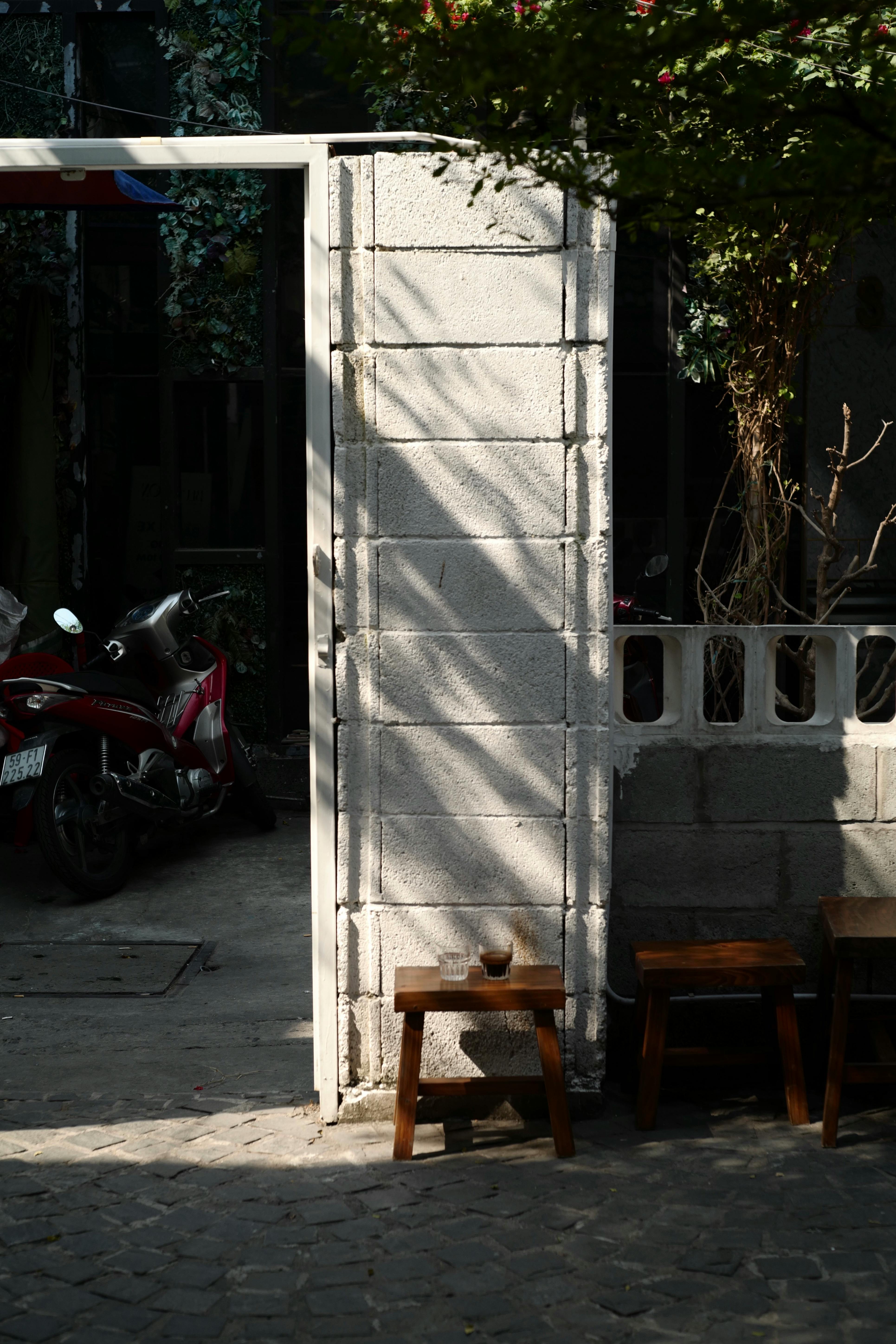 Sunlit urban alleyway with motorbike, stone wall, and wooden stools.