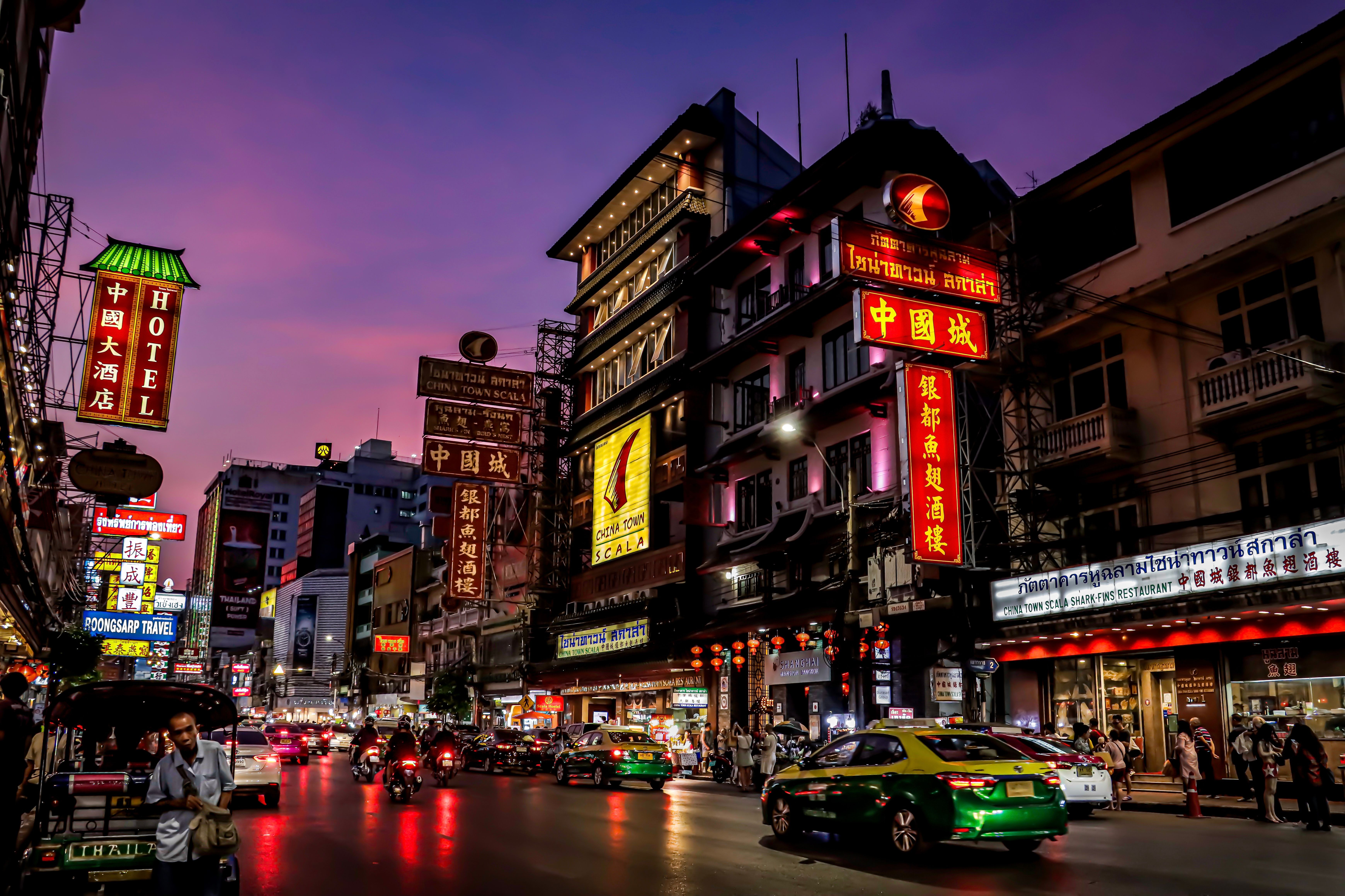 Colorful illuminated signs light up Bangkok's Chinatown street at dusk, showcasing urban life and culture.
