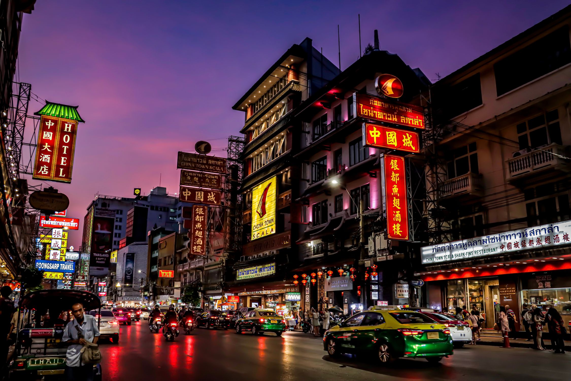 Colorful illuminated signs light up Bangkok's Chinatown street at dusk, showcasing urban life and culture.