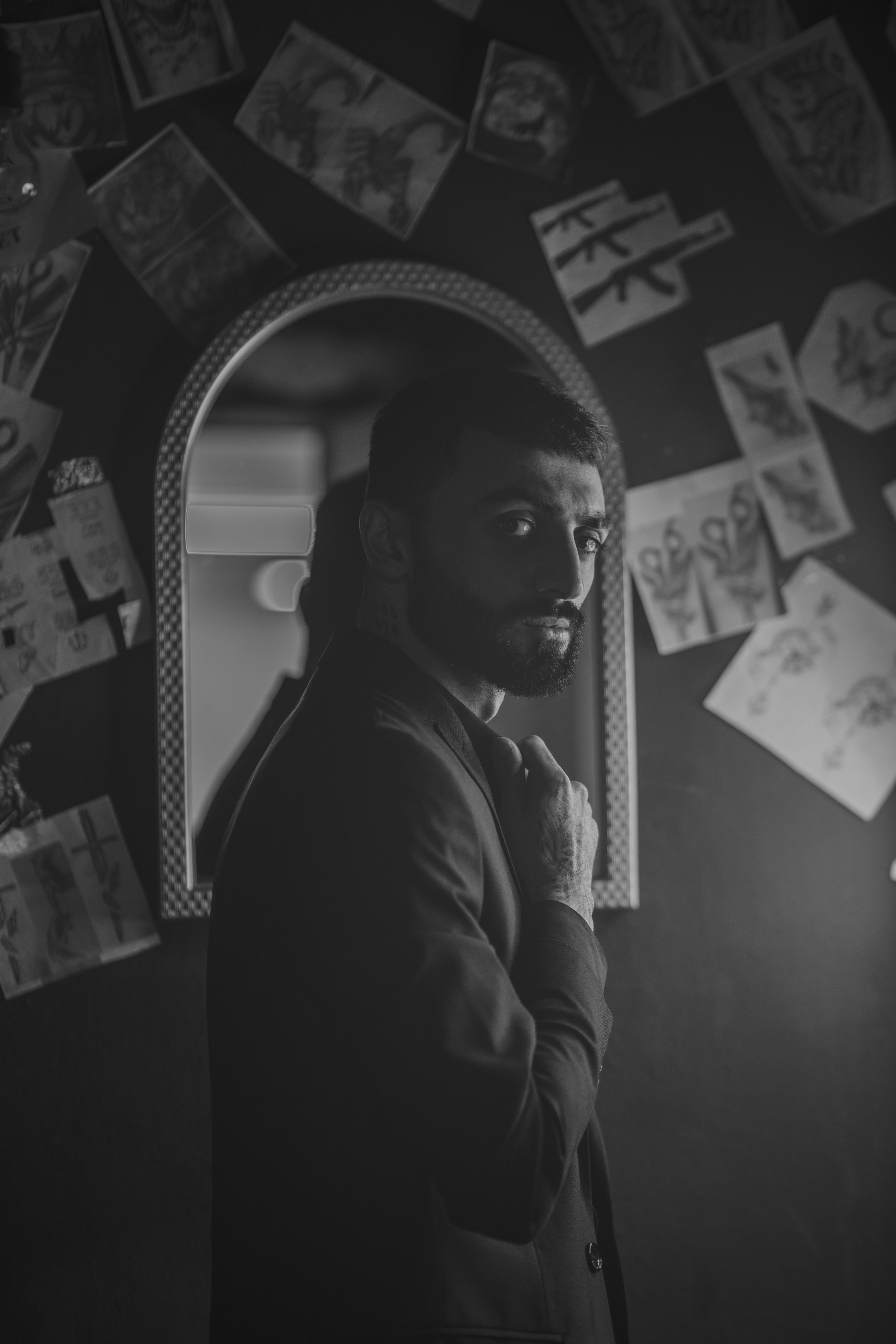 Dramatic black and white portrait of a man standing near a mirror, surrounded by sketches.