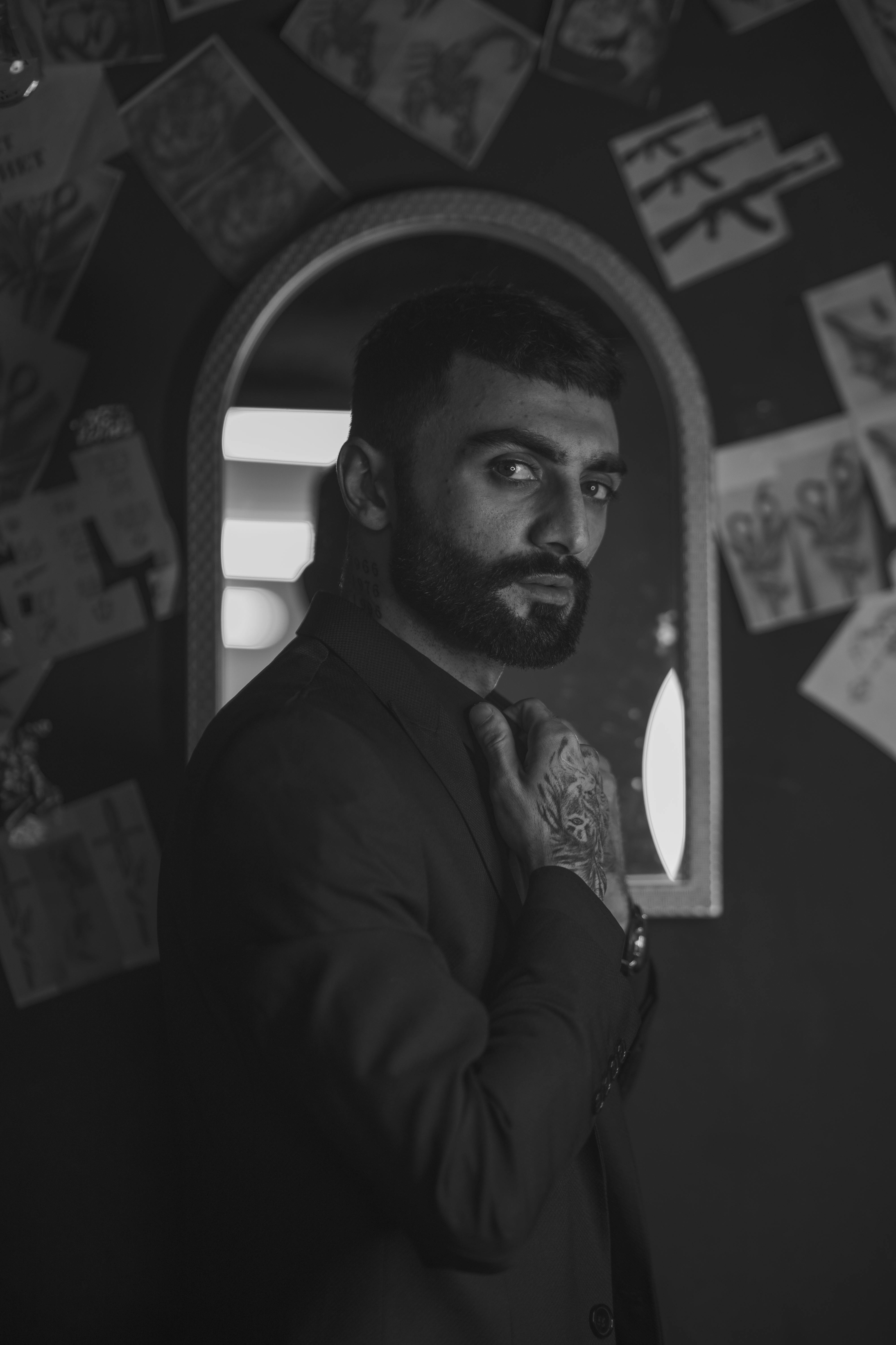 Black and white portrait of a man with a beard and tattoos in front of a mirror, wearing a black jacket.