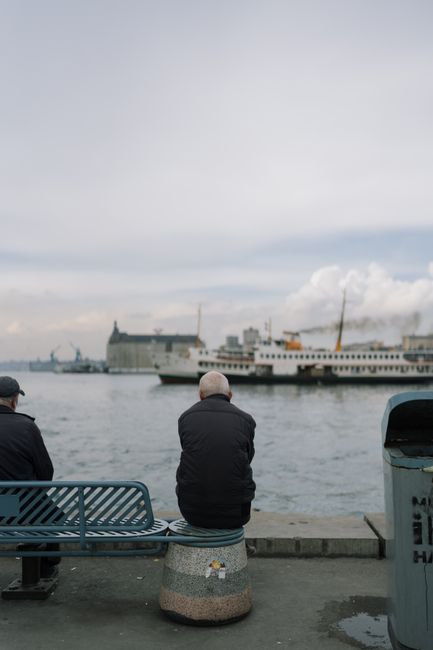 Elderly men sitting by the İstanbul harbor with ferries and skyline in view.