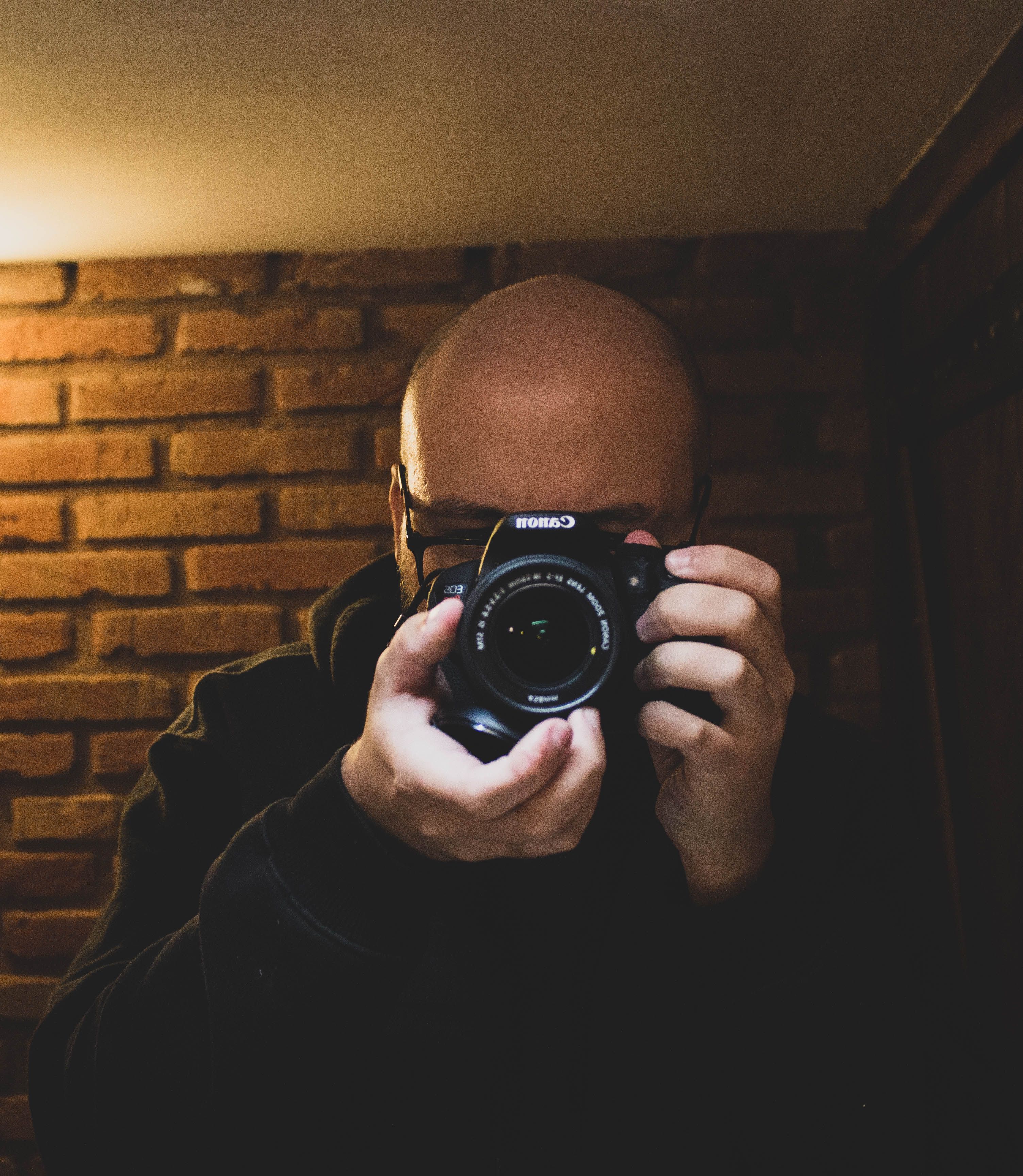 Bald man captures his reflection with a camera against a brick wall. Indoor setting in Florianópolis, Brazil.