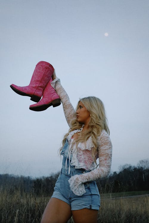 A woman in denim shorts holding up a pair of pink cowboy boots