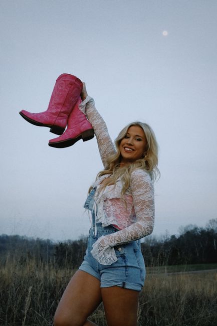 Smiling woman with long blonde hair raises pink cowboy boots in a rural field.