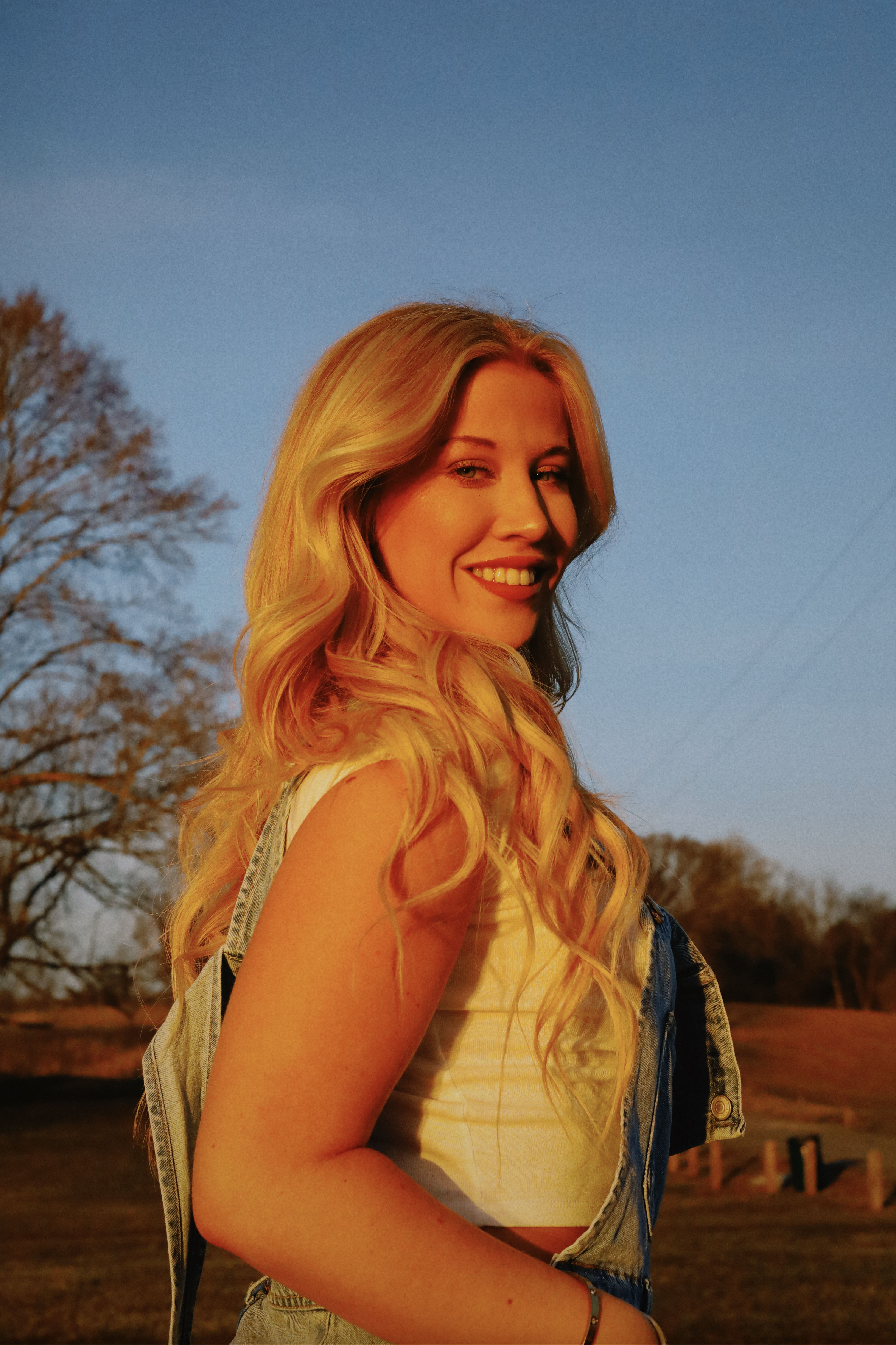 Sunlit portrait of a smiling woman in a tank top during golden hour, showcasing long blonde hair.