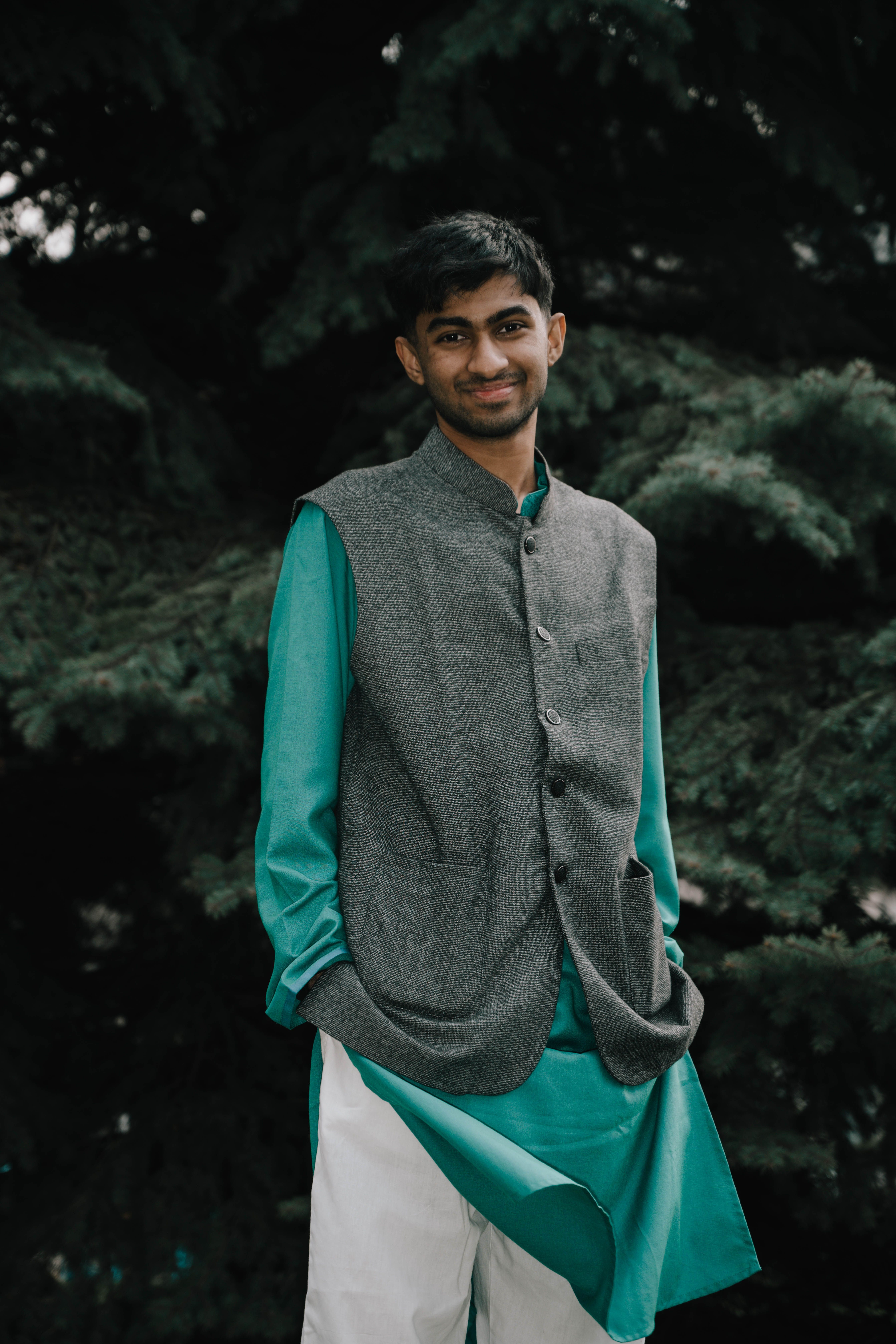 A smiling man wearing traditional Indian clothing stands against a backdrop of lush greenery.