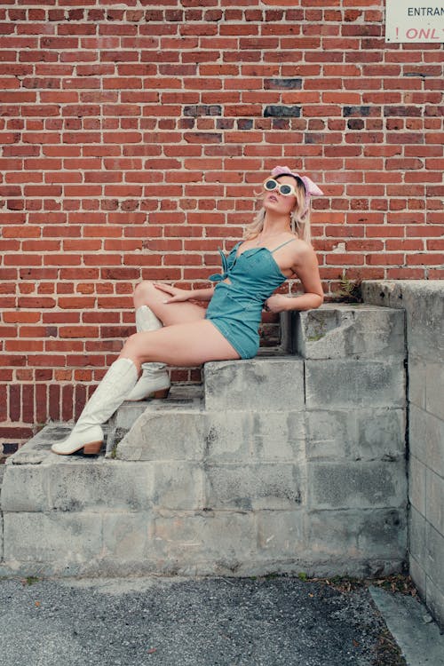 A woman in a blue dress and white boots sitting on a brick wall