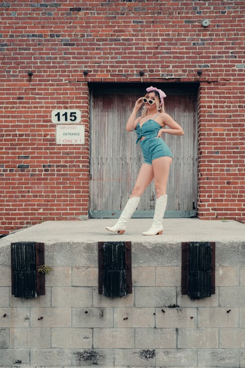 A woman in a blue dress and white boots posing on a brick wall