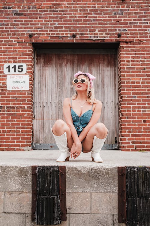 A blonde woman in a blue outfit and white boots sitting on a brick wall