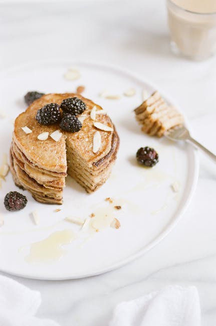 A high-angle shot of a stack of pancakes topped with blackberries and sliced almonds on a white plate.