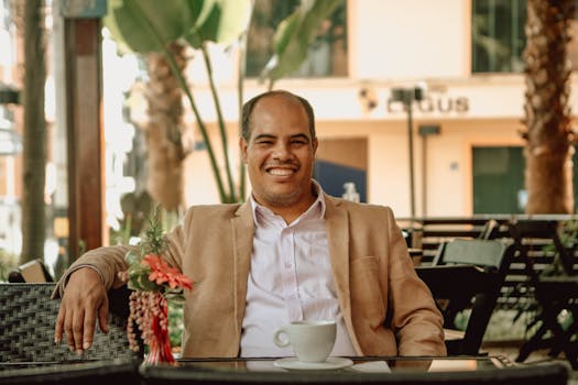 Balding man in a suit jacket smiling while enjoying a coffee in an outdoor café setting.
