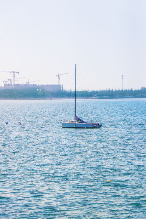 Free A tranquil scene of a yacht sailing on a calm ocean with a distant coastline and construction cranes visible under a clear summer sky. Stock Photo