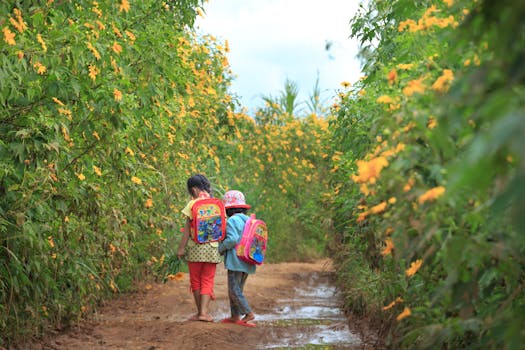 Two children with backpacks walking through a vibrant yellow flower path outdoors.