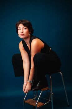 Stylish brunette woman in black outfit sitting on stool in studio.