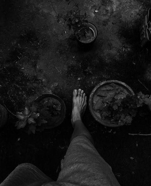 Monochrome image of a barefoot person standing near potted plants, creating a thoughtful scene.