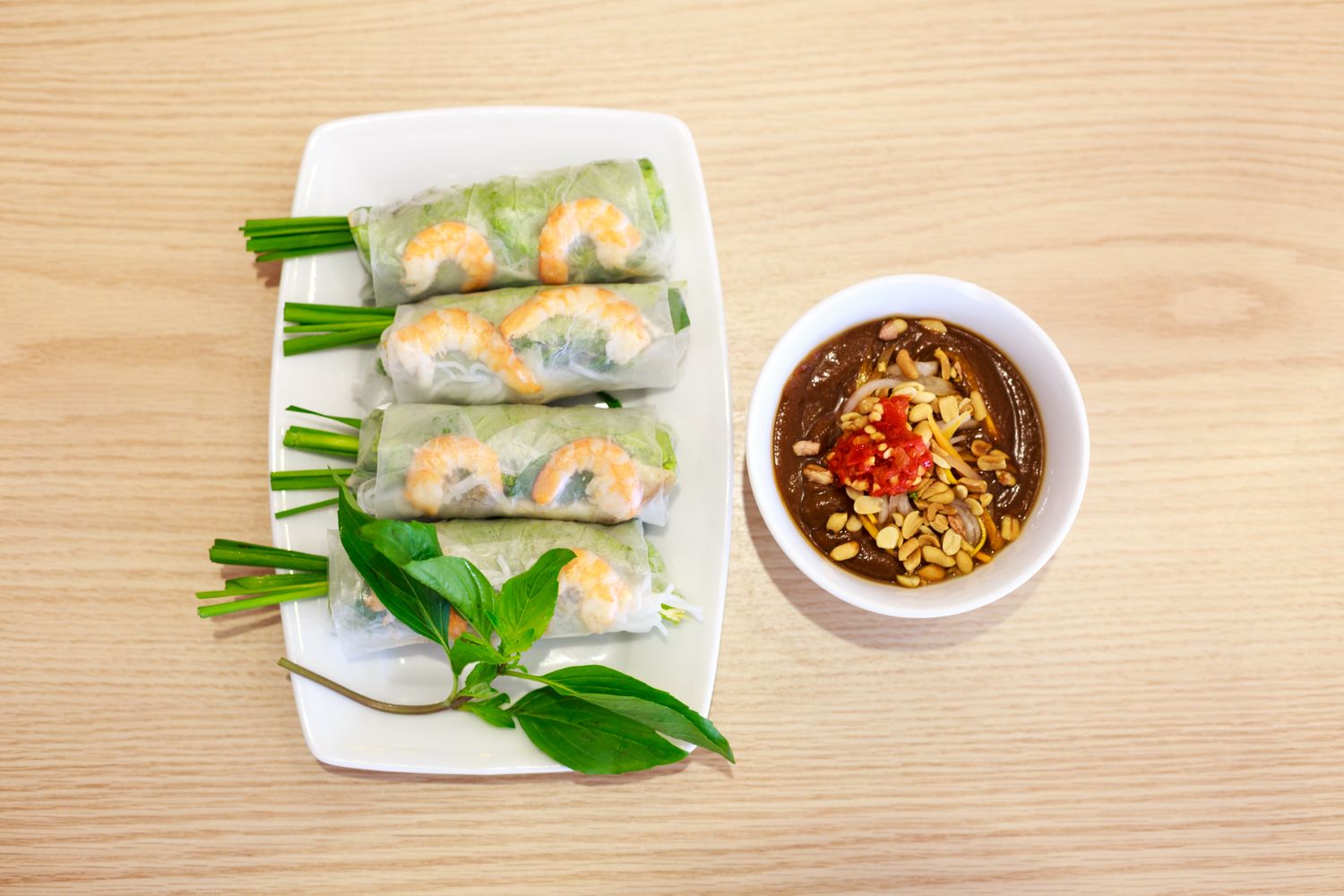 Four translucent Vietnamese summer rolls showing whole pink shrimp, rice noodles, leafy herbs and chive stems beside dark peanut-hoisin sauce topped with chopped peanuts and chile