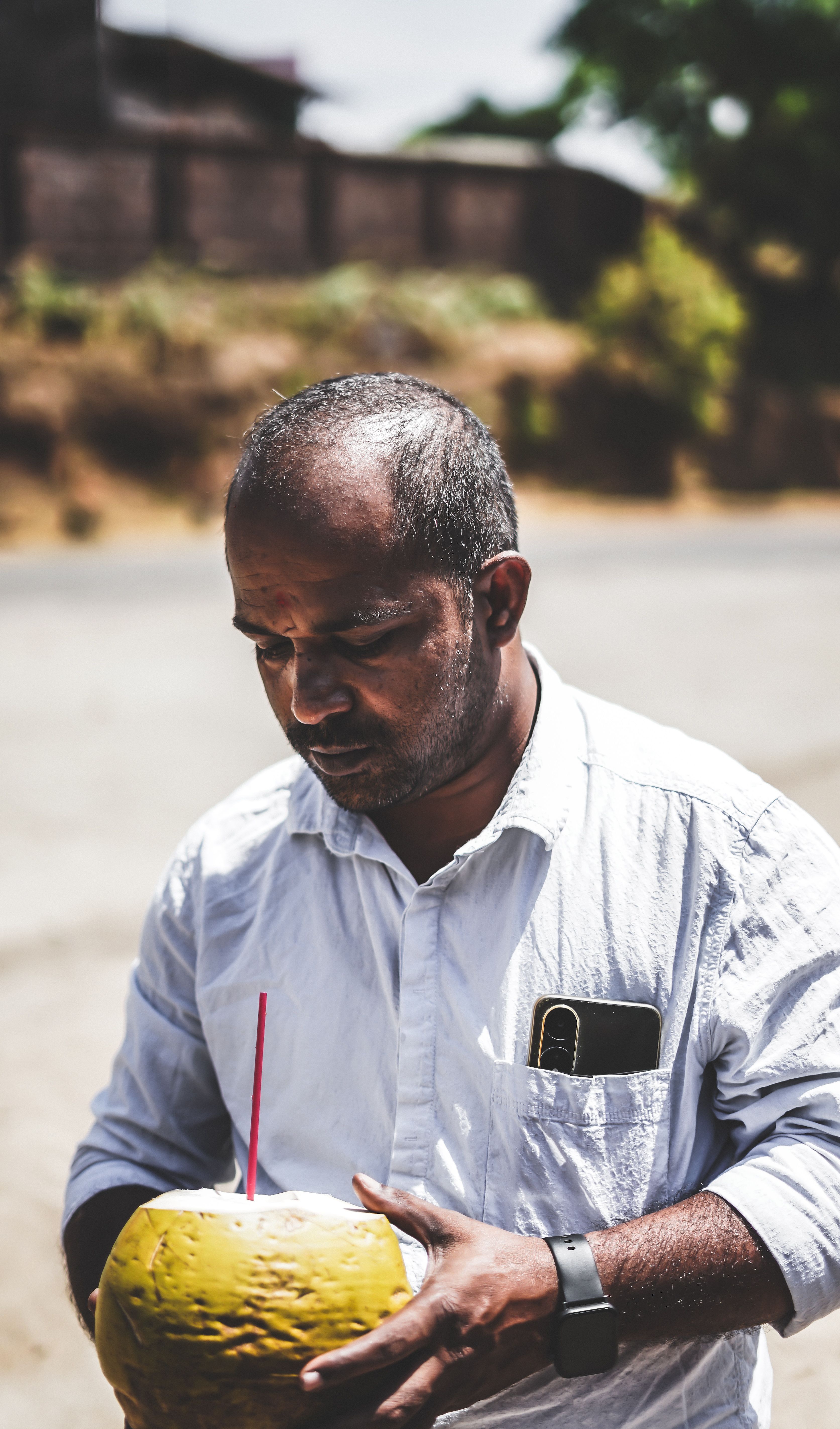 A middle-aged man in a white shirt drinks fresh coconut water with a pink straw outdoors on a sunny day.