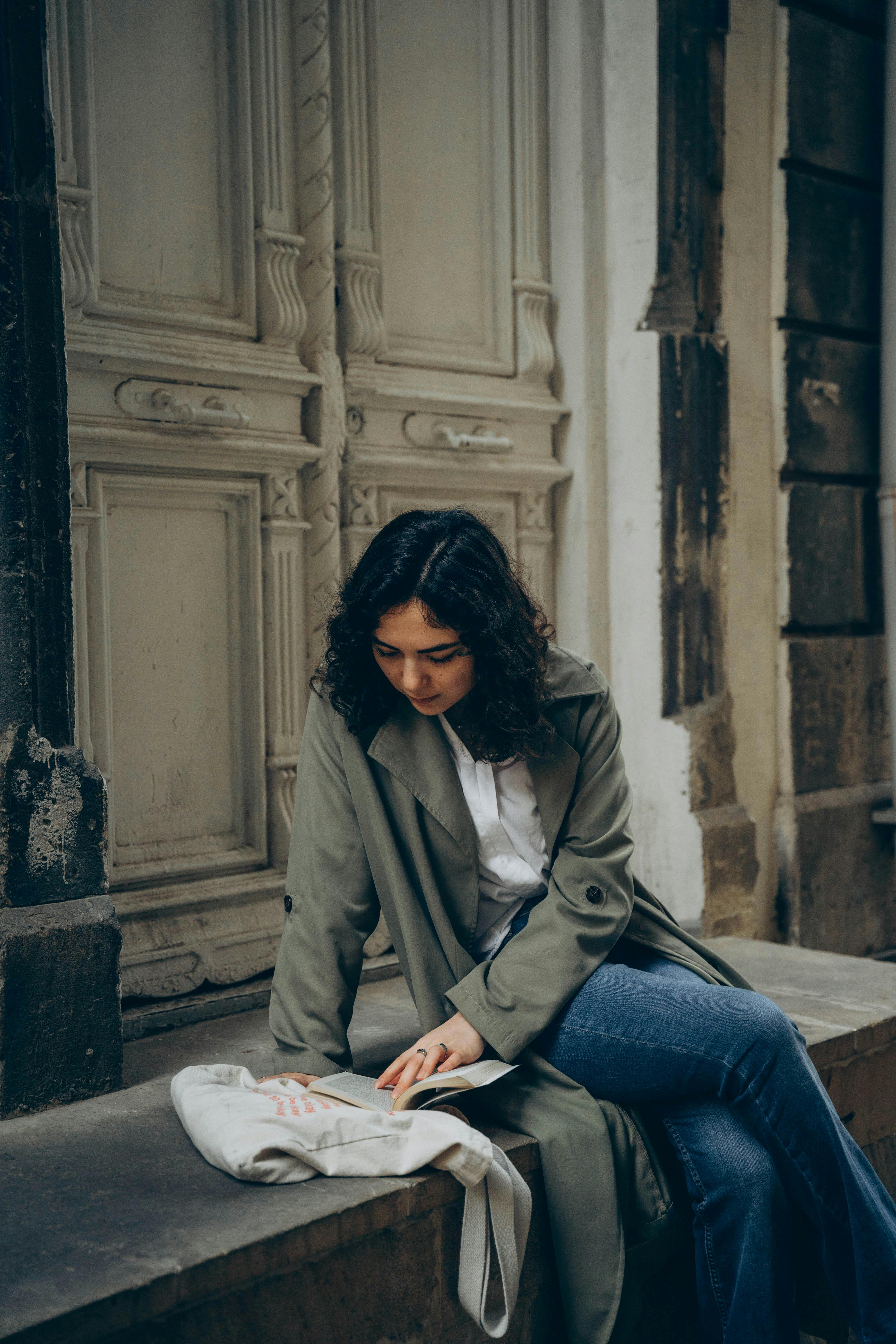 A young woman in a green coat sits by an old door, reading in an urban setting.