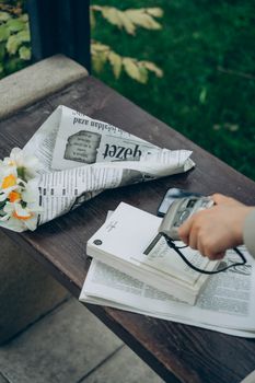 A person holding a camera over books and flowers on a bench outdoors.