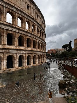 The Colosseum in Rome on a rainy day, with tourists exploring and dramatic skies.