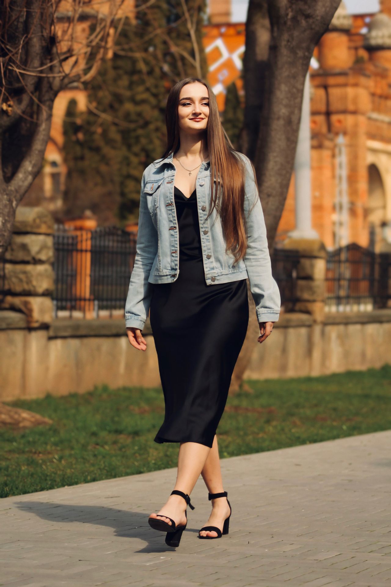 Woman wearing a black satin slip dress with a light-wash denim jacket and black block heel sandals