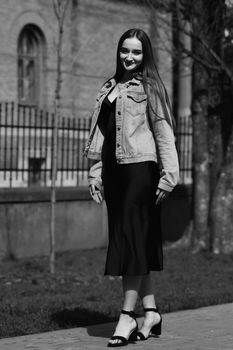 Elegant woman in black dress and denim jacket posing outdoors in monochrome.