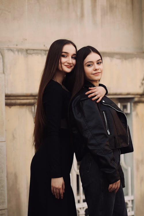 Free Young Woman in a Long Black Dress Embraces a Woman in a Leather Jacket and Black Jeans Stock Photo