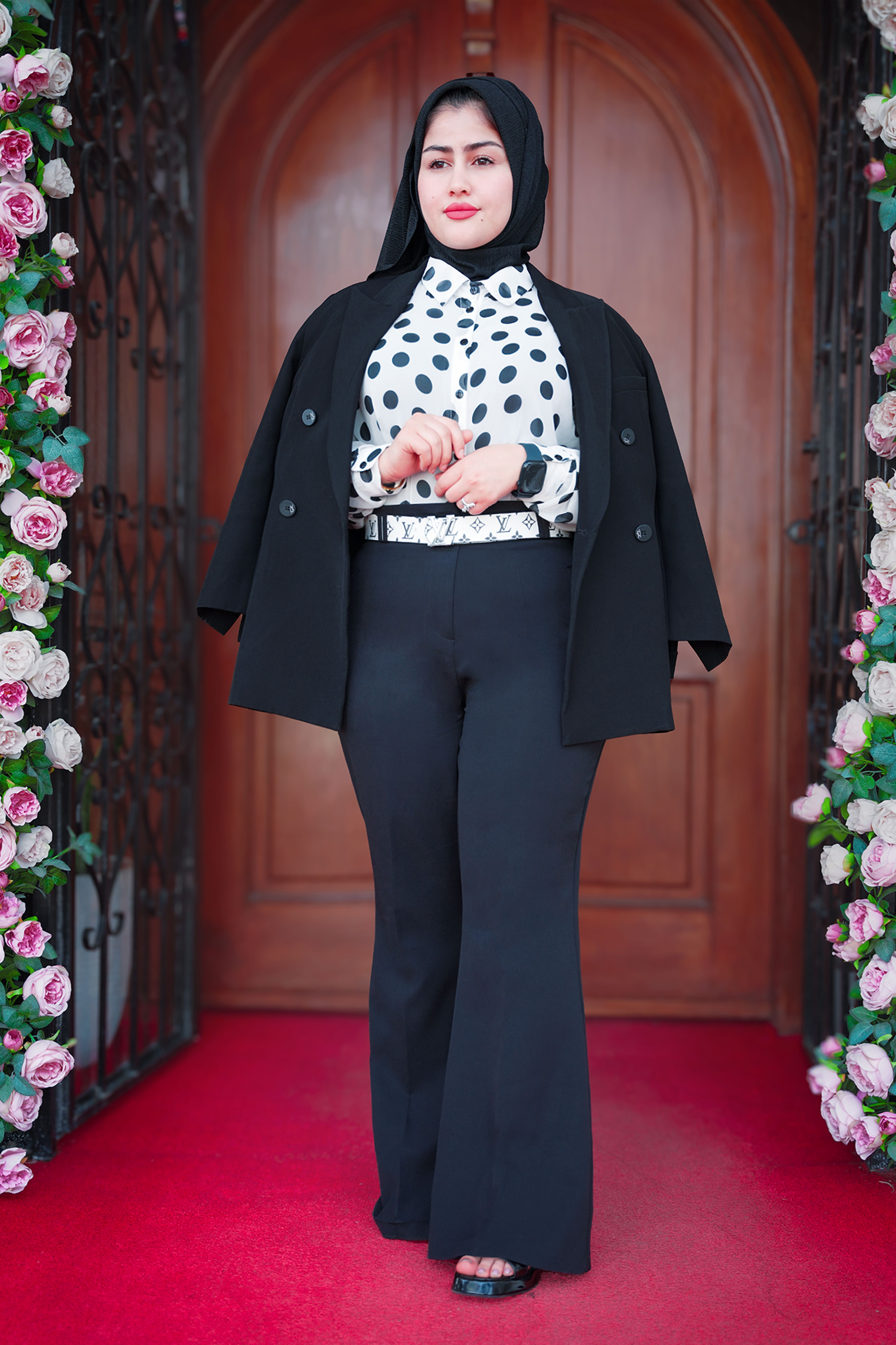Elegant woman in polka dot blouse and hijab poses confidently in front of wooden doors.