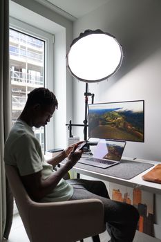 Adult man sits by the desk using phone, with laptop and monitor.