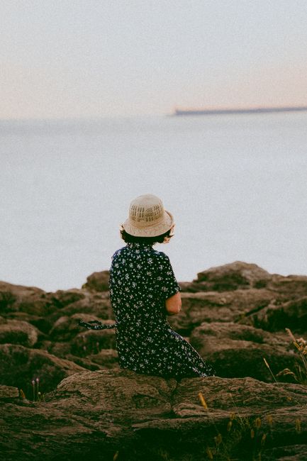 A woman in a sundress and hat sits on rocky seashore, gazing at the ocean.