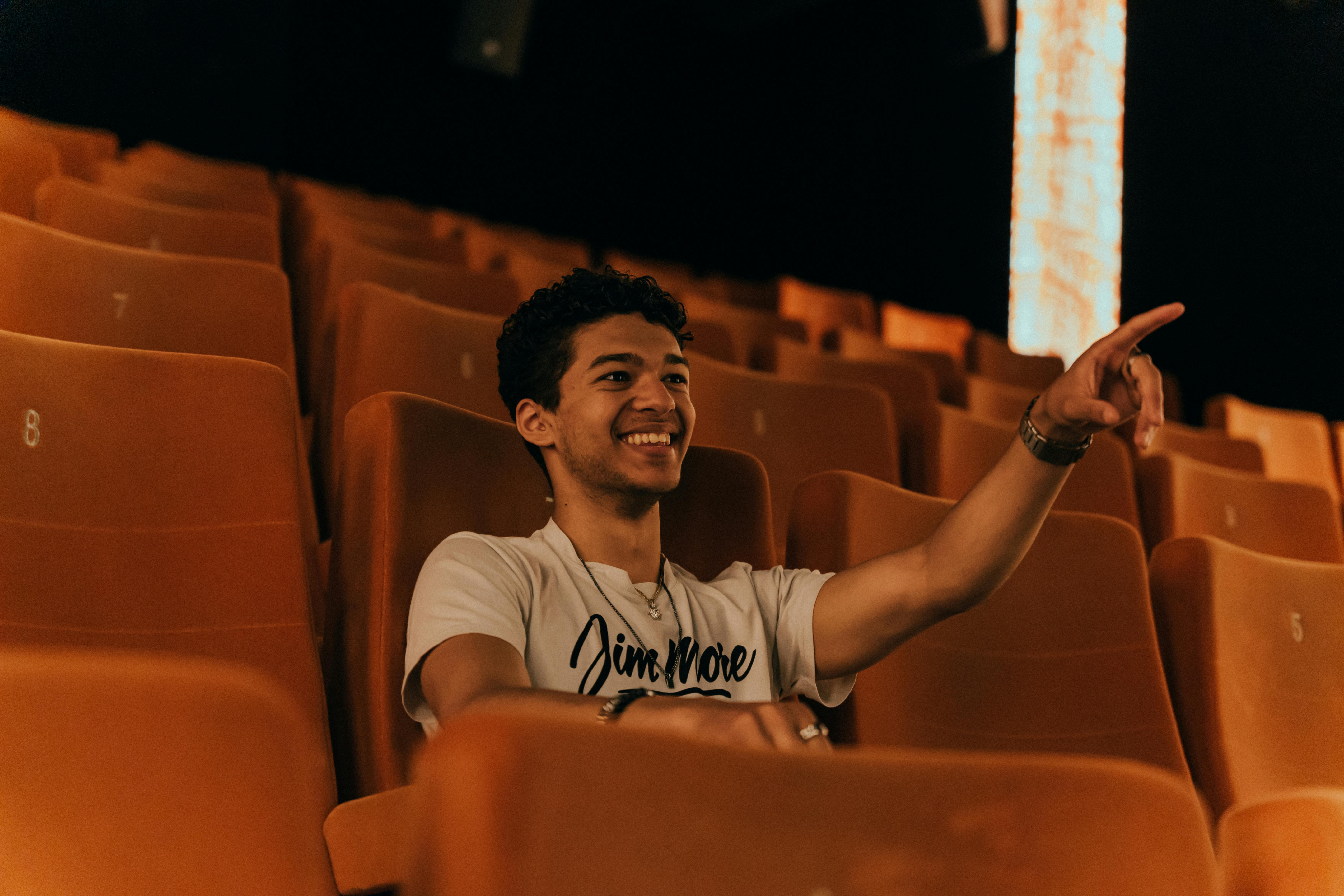 Free A young man with short hair smiles and points while seated in an empty cinema. Stock Photo